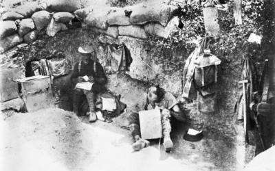 Two soldiers sitting in the 11th Battalion's Commanding Officer's dugout. The soldier on the right is sitting on the ground writing a letter using a box for support. 