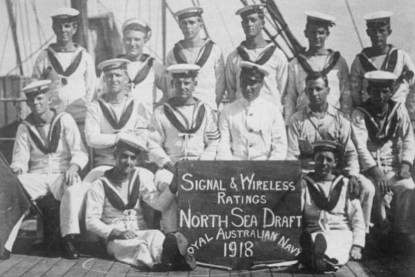Sailors sit behind a sign that reads "Signal & Wireless Ratings North Sea Draft, Royal Australian Navy, 1918