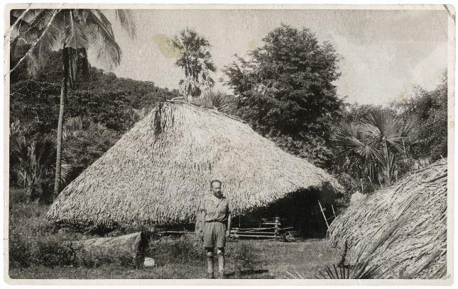 In the villague of Tuakau, Flying Officer Bill Arthur poses in front of two huts, dubbed “The Hyatt” and “The Hilton”.