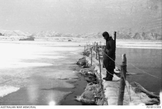 Korea, c. 1951-01. Carrying his rifle on his shoulder and wearing a greatcoat and pile cap against the winter cold, a member of the 3rd Battalion, The Royal Australian Regiment (3RAR), stands on a traditional Korean footbridge and stares into the frozen waters of the Han River. Beyond the soldier, the ice-bound river stretches away to a plain and hills covered in snow. 
