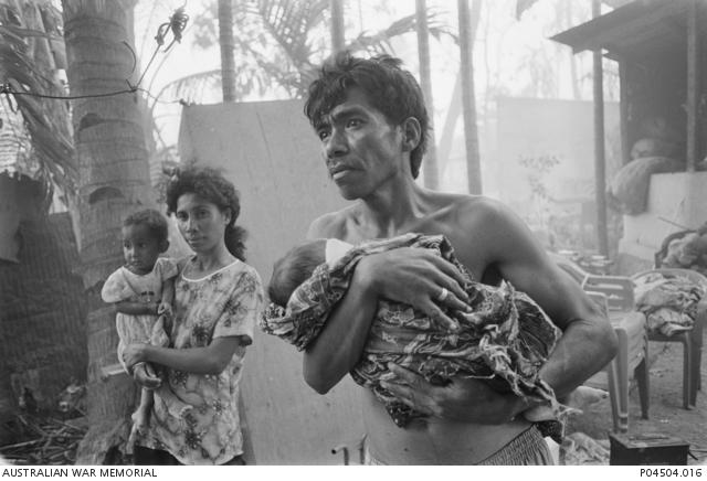 "David Dare Parker, Refugees recently returned to the burnt-out remains of their homes look on as more buildings burn, Dili, c.September 1999 P04504.016"