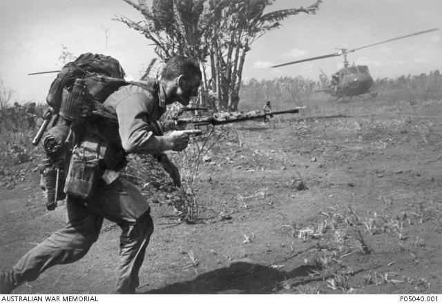 "An unidentified soldier of the 7th Battalion, The Royal Australian Regiment (7RAR), runs with a loaded pack and his rifle readied near Fire Support Base (FSB) Anne. A UH-1H 'Huey' Iroquois helicopter lands in the distance."