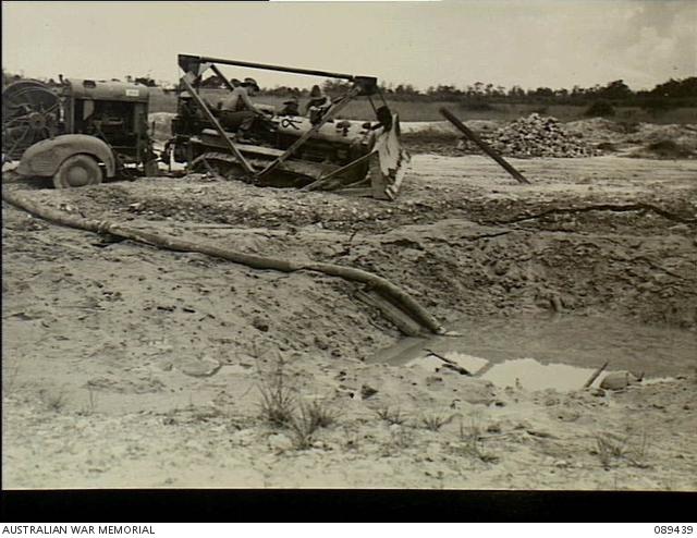 One of the numerous bomb craters on Tarakan airstrip being pumped dry to be filled by No 8 Airfield Construction Squadron, Royal Australian Air Force. The bogged tractor highlights the problems encountered during the operation.