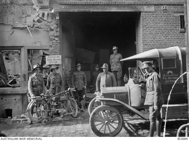 Men standing in the street with a motor vehicle in the foreground, one man standing with a motorbike, and a sign that reads "Australian war records section" hangs on the wall behind