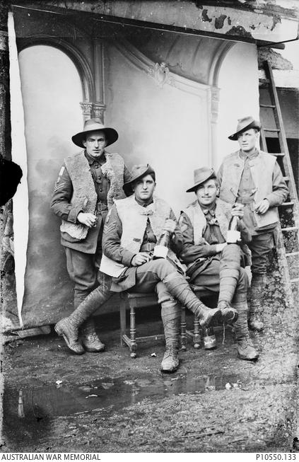 Group portrait of four members of the 13th Australian Light Trench Mortar Battery. The two soldiers in the front row hold bottles of French wine. Identified in this photograph are Corporal Robert Chaffey Stuart, third from left, and Private John Charles Bitton, fourth from left. The soldiers in this photograph each posed for their own individual portraits. In these portraits they are each wearing the same helmet, suggesting it was a prop owned by the photographers.